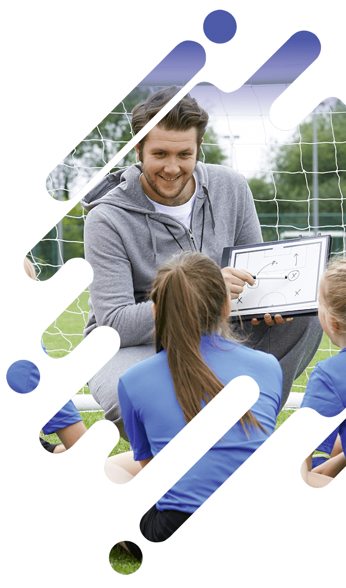 Ein Trainer im grauen Trainingsanzug zeigt jungen Sportlern in blauen Trikots auf einem Tablet eine Strategie. Sie sitzen zusammen auf einem Rasenplatz, im Hintergrund ist ein Fußballtor zu sehen.